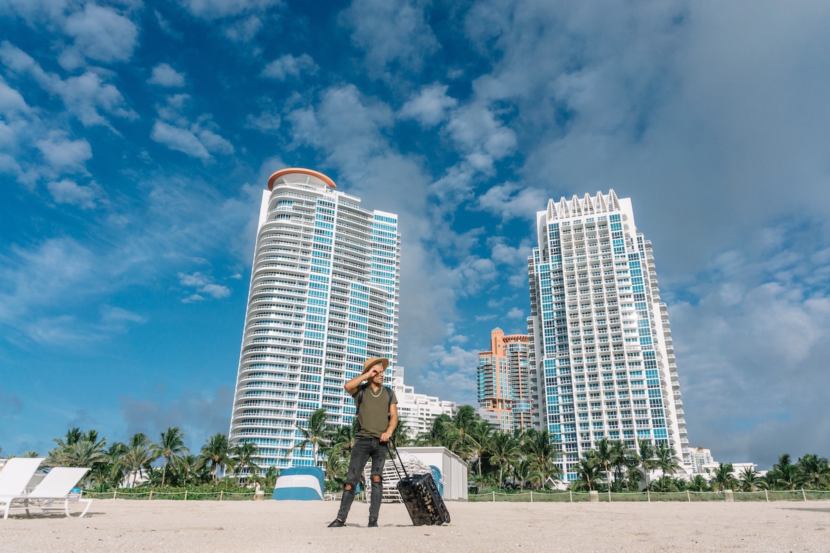 Traveler man for the first time in Miami. South beach buildings background. Handsome man with suitcase in hand on the white sand of South Miami Beach. SSUCv3H4sIAAAAAAAACpySz26DMAzG75P2DijnRgICBfYq0w7GCSVamlRJ6DRVffflD1Q574Z/jj9/tnm8v1UVmcFJJB/VI0Yhlkptzlvw0uiA69POrdBc2JLcLTvXJRBcemMlqBLO4HHVcBUB6k2piJ8pSZwHvznhYvcdIXhxCRoZviSyxc8cV0ciJUNFSBFyKpjb5sQOlMX/VZk/vo5p4CI0/ibDxSBWKAF5kN0j6Wa2TCNjFIZuoF0rOJ16PtCRIYO6EX3DIHXJ0uT7xwt7LVcBG5em2MLdIKj4gBWdb1ai1JeizPg1nekoQ7Npb6Pl16REGXODWcWTLEFTHHwF58JzfvCiEYafwlyLPtr4NPCuSni4XAwbxpqx72o2Tm3dDWNf7w/ytVcZdJKdQyjE+C15ObqMDgifYGjnhVGc64Z2536hI0JPcRId59hivyxhhc8/AAAA//8DALEgmWzIAgAA